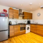 Cox & Co Estate agent in Edinburgh, Scotland. A modern kitchen with wooden cabinets, a silver refrigerator, a red microwave on top, a built-in oven with a stovetop, and a washing machine. Ideal for property investment or property management enthusiasts, it boasts black countertops, wooden floors, and a clock above the sink displaying 10:10.