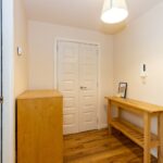 Cox & Co Estate agent in Edinburgh, Scotland. A well-lit, small entryway with a wooden floor, light beige walls, and a ceiling light. There's a wooden console table with a framed picture and an intercom on the wall above it, and a wooden cabinet next to a closed double door with decorative paneling—an ideal feature for property sales and lettings.