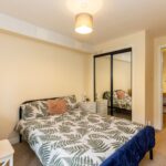 Cox & Co Estate agent in Edinburgh, Scotland. A cozy bedroom features a bed with a fern-patterned duvet cover, a small nightstand with a mirror, and a white desk. A mirrored closet reflects the bed, and a hallway with wooden flooring is visible. The room is well-lit with a ceiling light and table lamp—perfect for buyers interested in Property Sales and Lettings.