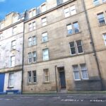 Cox & Co Estate agent in Edinburgh, Scotland. An image of a multi-story urban building with stone brick exterior, featuring large windows. The ground floor has a blue section to the left side. The street in the foreground has markings for "PERMIT HOLDERS." Ideal for an estate agent in Edinburgh specializing in property management. The sky is clear and blue.