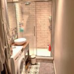 Cox & Co Estate agent in Edinburgh, Scotland. A narrow bathroom with a glass-enclosed shower featuring beige subway tile. A white sink sits atop a wooden vanity next to a washing machine. A grey rug is on the floor; a small trash bin is nearby. Towels hang on the left side, and toiletries are in the shower—ideal for property investment opportunities.
