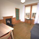 Cox & Co Estate agent in Edinburgh, Scotland. A simple living room with wooden flooring and white walls, ideal for property sales and lettings. It features a wooden dining table, chairs, a dark sofa, a fireplace with a wooden mantle, a small desk, and a built-in wooden shelf. Two large windows with sheer white curtains allow natural light to flood in.