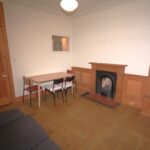 Cox & Co Estate agent in Edinburgh, Scotland. A modest living room with wooden flooring, featuring a small dining table with four chairs near a wooden door. There’s a fireplace with a wooden mantel on one wall, flanked by built-in shelves. A grey sofa is partially visible on the left side, offering potential for property management in Edinburgh.