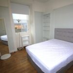 Cox & Co Estate agent in Edinburgh, Scotland. A small, minimalist bedroom with a bed featuring a tufted gray headboard and white bedding. The room, which any estate agent in Edinburgh would be proud to showcase, features wooden floors, a full-length mirror, a narrow bookshelf, a small nightstand with a lamp, and a window with a roller blind.
