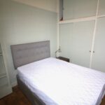 Cox & Co Estate agent in Edinburgh, Scotland. A small bedroom with a neatly made double bed featuring a white mattress cover. The bed has a grey padded headboard. There are built-in wardrobes with sliding doors in the background and built-in shelves on the left side. A small bedside table with a lamp is next to the bed—ideal for property investment in Edinburgh.