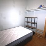 Cox & Co Estate agent in Edinburgh, Scotland. A small, sparsely furnished bedroom with a single mattress on a bed frame, a shelving unit with three shelves, and a wooden shoe rack. The light grey walls have patches, and there is a window on the right side letting in natural light—ideal for property investment opportunities.