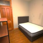 Cox & Co Estate agent in Edinburgh, Scotland. A small bedroom with a wooden floor features a single bed with a black headboard and white mattress, positioned against a light grey wall. Wooden shelves with black frames are on either side of the bed, and there is a wooden door with a window above it—ideal for property investment.