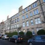 Cox & Co Estate agent in Edinburgh, Scotland. Image of a calm residential street lined with parked cars in front of a row of stone apartment buildings. The buildings, perfect for property investment, are several stories high and feature uniformly spaced windows. Scaffolding is visible on the leftmost building, indicating construction work.