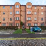 Cox & Co Estate agent in Edinburgh, Scotland. A four-story brick apartment building with multiple windows and three arched structures on the roof. Two cars are parked in front: a blue one on the right and a white one on the left. A grass lawn and a narrow walkway lead to the entrance—ideal for property management purposes.