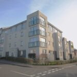 Cox & Co Estate agent in Edinburgh, Scotland. A modern four-story apartment building with a beige and gray exterior, featuring large windows and balconies, stands prominently on Ashwood Gait. A small hedge and grassy area grace the front, while a yellow car is parked on the nearby street corner under a clear and sunny sky.