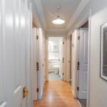 Cox & Co Estate agent in Edinburgh, Scotland. A brightly lit hallway with light wooden floors and white walls. Doors are seen on both sides, leading to various rooms of this property investment. Straight ahead is a bathroom with a visible toilet and sink. A cityscape photo hangs on the wall to the right under the ceiling light fixture illuminating the hallway.