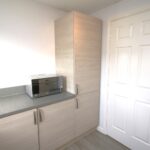 Cox & Co Estate agent in Edinburgh, Scotland. A modern kitchen with light wood cabinets and a gray countertop. A microwave oven is placed on the counter. A white door with a silver handle is to the right of the cabinets. The floor, in Ashwood Gait, is light-colored wood, blending well with the kitchen's minimalist design.