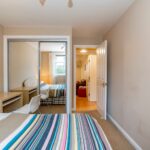 Cox & Co Estate agent in Edinburgh, Scotland. A bedroom with a striped bedspread, a wooden desk, and a white chair. There is a mirrored closet on the left and an open door leading to a brightly lit hallway. Another room with a bed and window is visible through the hallway. Beige walls complete this inviting setting, perfect for property investment.