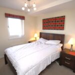 Cox & Co Estate agent in Edinburgh, Scotland. A cozy bedroom featuring a double bed with white bedding, flanked by two ashwood nightstands with lamps. A modern chandelier hangs from the ceiling, and a window with a red and black patterned valance allows natural light to enter. Matching artwork and a hat adorn the walls, adding a charming gait.