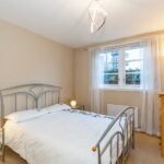 Cox & Co Estate agent in Edinburgh, Scotland. A bedroom with beige walls, featuring a metal bed frame, white bedding, and a nightstand with a lamp. There is a window with sheer white curtains, a tall wooden dresser, and a standing floor lamp. Ideal for property investment, the ceiling light has a modern, abstract design.