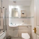 Cox & Co Estate agent in Edinburgh, Scotland. A small bathroom with white tiles and patterned trim features a bathtub with a glass shower screen, a white sink with vanity, cabinets, a mirror, and a toilet. The decor is minimalistic and clean, perfect for property sales and lettings, with all fixtures and fittings in white.