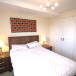 Cox & Co Estate agent in Edinburgh, Scotland. A clean, minimalist bedroom in Ashwood Gait features a double bed with a wooden headboard and white bedding. Two bedside tables hold lamps, while a red and grey abstract painting hangs above the bed. A white, double-door closet is on the right, and a modern ceiling light fixture completes the space.
