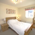 Cox & Co Estate agent in Edinburgh, Scotland. A cozy bedroom at Ashwood Gait with a wooden bed frame and white bedding. The room features a matching wooden nightstand, a small lamp, and a full-length mirror. The light-colored walls are adorned with three abstract art pieces above the bed, enhancing the space’s charm. A window with patterned curtains completes the look.