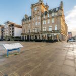 Cox & Co Estate agent in Edinburgh, Scotland. A historic building with a clock tower stands on a cobblestone street, surrounded by modern buildings. In the foreground, there is an information board with text and illustrations. Several cars are parked along the street. The sky is partly cloudy—a perfect scene for an estate agent in Edinburgh to showcase property sales and lettings.