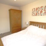 Cox & Co Estate agent in Edinburgh, Scotland. A serene bedroom features an ashwood bed with white bedding and two wicker bedside tables adorned with white lamps. A wooden wardrobe stands against the wall, and above the bed hangs a colorful, abstract patterned canvas. The walls are painted in a light cream color, setting a tranquil gait.