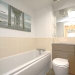 Cox & Co Estate agent in Edinburgh, Scotland. A modern bathroom with light beige tiled walls and a white bathtub. A sink with an ashwood cabinet and a toilet are on the right side. Above the sink is a large mirror, and above the bathtub hangs a painting of a pier extending into the ocean.