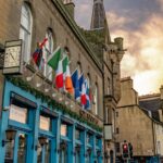 Cox & Co Estate agent in Edinburgh, Scotland. Street view of a building adorned with international flags, including those of Italy, Ireland, France, and the Netherlands. The building with large blue-framed windows, a vintage clock, and a "Fransiscan" sign stands as an example of excellent property management. A church spire and historic buildings are in the background.