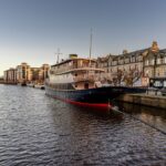 Cox & Co Estate agent in Edinburgh, Scotland. A boat is moored at a dock in a canal lined with historical buildings and modern apartments, perfect for property investment. The sky is clear and blue, and the water is calm. Parked cars are visible along the right side, with additional boats and structures in the background.