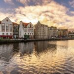 Cox & Co Estate agent in Edinburgh, Scotland. A serene waterfront scene at sunset. Several historic buildings, with varied architectural styles, line the body of water, their reflections appearing in the still water. The sky is filled with dramatic clouds, colored warmly by the setting sun—an ideal backdrop for any estate agent in Edinburgh showcasing property sales and lettings.