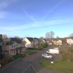 Cox & Co Estate agent in Edinburgh, Scotland. Ashwood Gait is a suburban residential street with modern houses and cars parked along the road. The sky is clear and blue, and there is green grass in the yards and common areas. Leafless trees in the background indicate it might be late fall or winter.