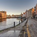 Cox & Co Estate agent in Edinburgh, Scotland. A cobblestone street runs alongside a tranquil river, lined with historic buildings in warm, earthy tones. This charming setting, ideal for property sales and lettings, is bordered by black posts and chains, with two yellow lines marking the road. The sky is clear and blue, suggesting a calm and peaceful day.