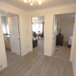 Cox & Co Estate agent in Edinburgh, Scotland. A hallway with light wooden flooring and beige walls at Ashwood Gait. Three open doorways are visible: one leads to a modern kitchen, another to a living room with a dining area, and the third to a bedroom. There is a mirror on one wall and a light fixture on the ceiling.