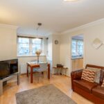 Cox & Co Estate agent in Edinburgh, Scotland. A cozy living room features a brown leather sofa with patterned cushions, a beige rug, and a TV on a light wooden stand. In the adjacent dining area, there's a table with four chairs and a window. The kitchen is visible through an open doorway. Perfect for those considering property investment, decor includes wall art and a clock.