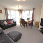 Cox & Co Estate agent in Edinburgh, Scotland. A spacious living room with beige walls and grey carpet. It features a black leather sofa set with red and white cushions, a stylish Ashwood dining table by the window, and a flat-screen TV on a stand. Large windows with striped curtains allow ample natural light.