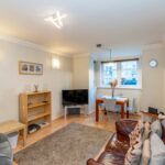 Cox & Co Estate agent in Edinburgh, Scotland. A cozy living room features a brown leather couch with pillows, an armchair, and a TV on a stand beneath shelves. A small dining area with a round table and four chairs sits by a window. Modern light fixtures and minimalistic decor give the room a warm and welcoming feel, ideal for property investment.