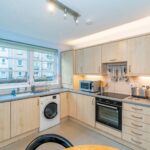 Cox & Co Estate agent in Edinburgh, Scotland. A modern kitchen with light wooden cabinets and a U-shaped countertop. It features a washing machine, microwave, oven, stovetop with black hood, stainless steel sink, and various utensils hanging on the wall. There's a large window with a green roller blind; ideal for Property Sales and Lettings.