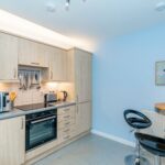 Cox & Co Estate agent in Edinburgh, Scotland. A modern kitchen with light wood cabinetry, an electric stove, oven, microwave, and utensils hanging above the counter. A circular wooden table is accompanied by two black bar stools, and a framed abstract artwork adorns the light blue wall—perfect for property sales and lettings.