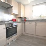 Cox & Co Estate agent in Edinburgh, Scotland. A modern kitchen with Ashwood cabinets, gray countertops, and stainless steel appliances, including a built-in oven and extractor hood. A window above the sink allows natural light to enter. Small appliances like a kettle and toaster adorn the counter.