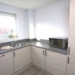 Cox & Co Estate agent in Edinburgh, Scotland. A modern kitchen at Ashwood Gait features light-colored wooden cabinets, a gray countertop, a round cutting board beside a sink under a window, a microwave next to the cabinets, and some potted plants on the windowsill. The floor is light gray wood.
