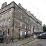 Cox & Co Estate agent in Edinburgh, Scotland. A view of Dundonald Street shows a residential block with four-story stone buildings along a narrow, slightly curved road. Several cars are parked along the curb. The buildings have tall windows, chimneys, and wrought-iron fences. The sky is cloudy, and the area appears quiet.