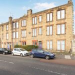 Cox & Co Estate agent in Edinburgh, Scotland. A row of three-story, stone-built apartment buildings on Piersfield Terrace in Edinburgh, EH8, with several cars parked along the street in front. The buildings have large windows and some orange and gray doors. A blue sky with scattered clouds is visible above.