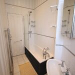 Cox & Co Estate agent in Edinburgh, Scotland. A narrow bathroom on Dundonald Street features a white tiled bathtub with a black base, a mounted showerhead, and a glass shower screen. Next to the bathtub is a white sink with dual faucets and a mirror cabinet above. A yellow mat brightens the tiled floor near the door.