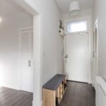 Cox & Co Estate agent in Edinburgh, Scotland. A modern, clean hallway at Piersfield Terrace, Edinburgh, EH8 features a white front door with a frosted window, a small bench with storage cubbies underneath, and a radiator along one wall. A closet with louvered doors is partially visible in the adjacent room. The walls are a pale gray.
