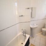 Cox & Co Estate agent in Edinburgh, Scotland. A small, minimalist bathroom on Dundonald Street features white square tiles on walls and floor. Includes a bathtub with chrome fixtures, a wall-mounted sink with a mirror above, a toilet, and a heated towel rail. Natural light enters through a window near the ceiling.