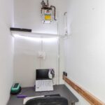 Cox & Co Estate agent in Edinburgh, Scotland. A small workspace in a narrow room on Piersfield Terrace, EH8, features a grey desk with a black mesh chair. The desk holds a laptop with a white keyboard, a green pencil case, and a purple pen. A gas meter and pipe are mounted on the wall above an LED strip light in this Edinburgh setup.