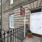 Cox & Co Estate agent in Edinburgh, Scotland. A stone building on Dundonald Street with a white door numbered 24 displays a "To Let" sign from Cox & Co. The entrance is accessed by a black metal railing along the pathway. There's a tree visible in the background and a potted plant near the door.