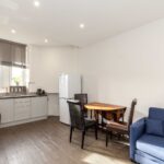 Cox & Co Estate agent in Edinburgh, Scotland. A modern, well-lit kitchen and living area in Piersfield Terrace, Edinburgh, featuring a white refrigerator, small round wooden dining table with two chairs, L-shaped white kitchen cabinets, stainless steel appliances, and a blue sofa. A window with gray blinds lets in natural light, illuminating the EH8 space.