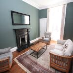 Cox & Co Estate agent in Edinburgh, Scotland. A cozy living room on Dundonald Street with green walls features a black fireplace with a large mirror above it. The room has a beige sofa, two matching armchairs, a wooden coffee table, and a patterned rug. Natural light streams in from a large window with beige curtains.