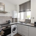 Cox & Co Estate agent in Edinburgh, Scotland. Modern kitchen in Piersfield Terrace with white walls and gray cabinets. Includes a stove, oven, washing machine, kettle, coffee maker, and small plant by the window with gray blinds. Countertop is light gray, backsplash is tiled, and an overhead range hood is present above the stove in this EH8 property.
