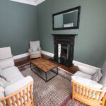 Cox & Co Estate agent in Edinburgh, Scotland. A cozy living room on Dundonald Street with green walls, featuring a black fireplace with a mirror above it. The room includes a white sofa, two matching armchairs, and a wooden coffee table on a patterned rug. Radiator and hardwood floors are visible.