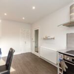 Cox & Co Estate agent in Edinburgh, Scotland. A modern kitchen and dining area on Piersfield Terrace, Edinburgh, featuring a black electric stove with an oven, a stainless steel range hood, white walls, dark wooden flooring, and recessed lighting. To the left is a small wooden table with black and brown chairs. A cupboard and shelves line the walls in EH8.